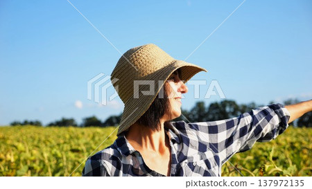 Happy smiling female farmer looks into sunflower field and enjoys harvest. Portrait of adult beautiful agronomist in straw hat with yellow plantation at background. Concept of agricultural business Happy smiling female farmer looks into sunflower field and enjoys harvest. Portrait of adult beautiful agronomist in straw hat with yellow plantation at background. Concept of agricultural business 137972135