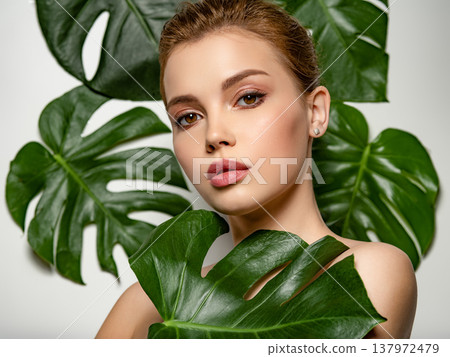 Closeup face of young beautiful woman with a healthy clean skin. Beautiful white girl with big green leaves. Beauty and spa treatment concept. Pretty woman with natural makeup and plant near face Closeup face of young beautiful woman with a healthy clean skin. Beautiful white girl with big green leaves. Beauty and spa treatment concept. Pretty woman with natural makeup and plant near face 137972479