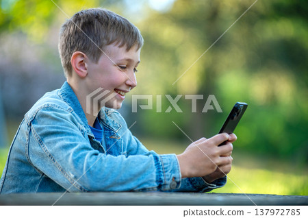Happy teen boy playing on a cell phone outdoors. Young cheerful teen typing on mobile phone sitting behind a table in a park. Smiling child is texting on the phone in a garden, on a sunny summer day. Happy teen boy playing on a cell phone outdoors. Young cheerful teen typing on mobile phone sitting behind a table in a park. Smiling child is texting on the phone in a garden, on a sunny summer day. 137972785