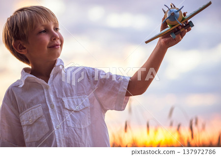 Happy child playing with a toy plane in nature during summer sunset. Boy in a white shirt with a plane in hands on wheat field. Kid holds a wooden airplane and dreams of being a pilot, on the nature Happy child playing with a toy plane in nature during summer sunset. Boy in a white shirt with a plane in hands on wheat field. Kid holds a wooden airplane and dreams of being a pilot, on the nature 137972786