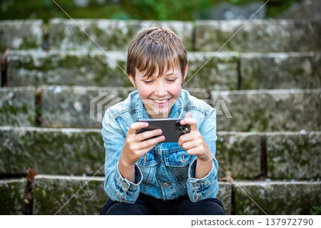A happy young kid is sitting outside with a phone. Smiling teen boy outdoors with a smartphone in his hands. 137972790