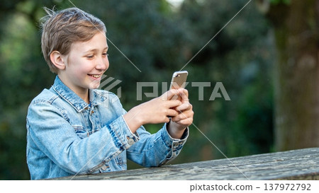 A happy young kid is sitting outside with a phone. Smiling teen boy outdoors with a smartphone in his hands. A happy young kid is sitting outside with a phone. Smiling teen boy outdoors with a smartphone in his hands. 137972792
