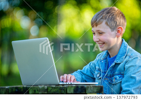 Happy Student with a laptop sits behind a table in a park. School boy is typing on computer outdoors. Cheerful young boy studying online with a computer, outdoors. Boy on online lessons while outside. Happy Student with a laptop sits behind a table in a park. School boy is typing on computer outdoors. Cheerful young boy studying online with a computer, outdoors. Boy on online lessons while outside. 137972793