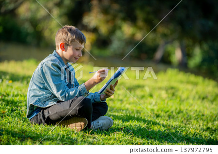 Happy student with a textbook in his hands is sitting on a green lawn in a park. A thinking student writing in a notebook in a garden. Young boy reading his notes and memorizing the text before a Happy student with a textbook in his hands is sitting on a green lawn in a park. A thinking student writing in a notebook in a garden. Young boy reading his notes and memorizing the text before a 137972795