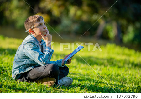 Thinking student with a textbook in his hands is sitting on a green lawn in a park. Young boy reading his notes and memorizing the text before a test. A student writing in a notebook in a garden Thinking student with a textbook in his hands is sitting on a green lawn in a park. Young boy reading his notes and memorizing the text before a test. A student writing in a notebook in a garden 137972796