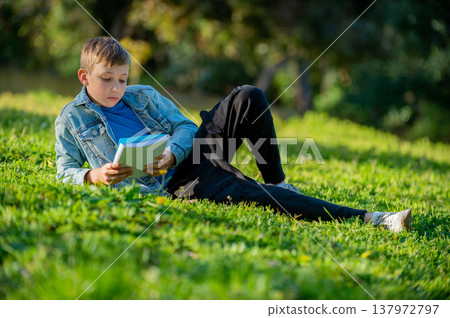 Schoolboy with a textbook in his hands is lying on a green lawn in a park during summer day. Studying teen looks to the notebook in a garden. Pupil reading his notes and memorizing the text outdoors Schoolboy with a textbook in his hands is lying on a green lawn in a park during summer day. Studying teen looks to the notebook in a garden. Pupil reading his notes and memorizing the text outdoors 137972797