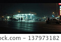 A gas station at night with illuminated pumps. Several cars are parked, and a few people are standing near the entrance. The scene is foggy and urban. 137973182