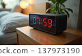 Digital alarm clock on a wooden nightstand in a cozy modern bedroom with a green plant 137973190