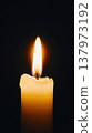 A lit candle with a warm flame against a dark background. The wax is partially melted, creating a soft glow around the flame. 137973192