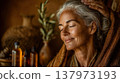An elderly woman with gray hair enjoys a relaxing spa treatment. Natural oils and pottery are visible in the background, creating a serene atmosphere. 137973193