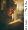 Young Woman Reading Paperback Book by Window in Warm Golden Hour Light 137974167