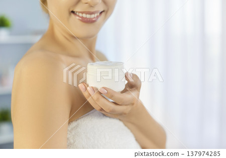 Pretty smiling young woman holding white jar with moisturizing body or face cream standing at home Pretty smiling young woman holding white jar with moisturizing body or face cream standing at home 137974285