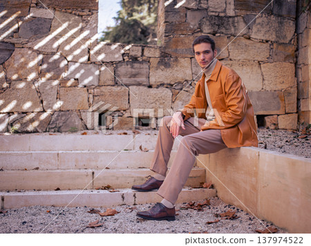 Spring fashion. Handsome male model in a brown leather jacket, beige trousers and sweater posing in an antique outdoor setting. Young man wearing stylish autumn clothes in brown and beige tones. Spring fashion. Handsome male model in a brown leather jacket, beige trousers and sweater posing in an antique outdoor setting. Young man wearing stylish autumn clothes in brown and beige tones. 137974522
