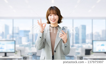 一位微笑的女人/女商人拿著智慧型手機,做出「OK」的姿勢 一位微笑的女人/女商人拿著智慧型手機,做出「OK」的姿勢 137975997