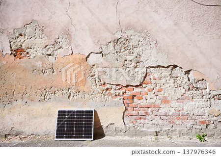 Photovoltaic solar panel at weathered, textured wall with peeling paint. Juxtaposition of modern renewable energy technology against rustic, decaying backdrop highlights contrast between old and new. 137976346