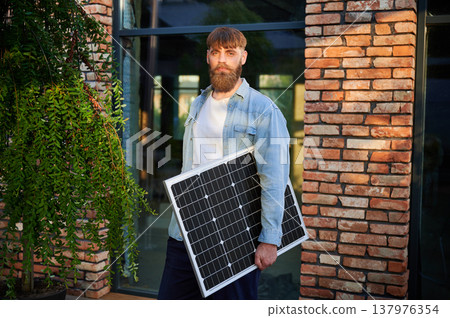 Portrait of bearded man holds solar panel, standing in front of brick building and lush greenery. Guy in shirt and t-shirt exudes calm and eco-conscious vibe, blending nature with urban elements. 137976354