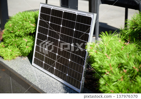 Photovoltaic solar panel against backdrop of vibrant green shrubs on stone ledge, soaking up sunlight in modern, urban landscape. Concept of integration of renewable energy within city environments. Photovoltaic solar panel against backdrop of vibrant green shrubs on stone ledge, soaking up sunlight in modern, urban landscape. Concept of integration of renewable energy within city environments. 137976370