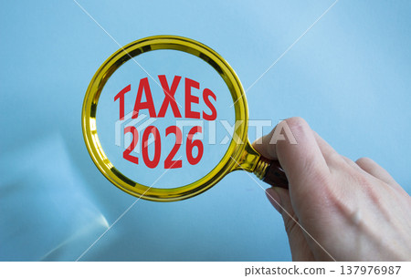 TAXES 2026 symbol. Magnifying Glass with words TAXES 2026. Businessman hand. Beautiful blue background. Business and TAXES 2026 concept. Copy space. 137976987