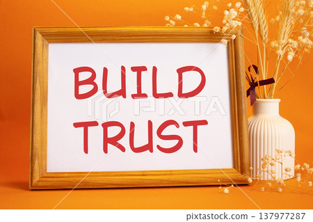 Build trust symbol. Concept words Build trust on beautiful wooden picture frame. Beautiful orange background with flower vase. Business and Build trust concept. Copy space. Build trust symbol. Concept words Build trust on beautiful wooden picture frame. Beautiful orange background with flower vase. Business and Build trust concept. Copy space. 137977287
