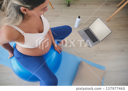 Pregnant woman exercising on fitness ball watching laptop Pregnant woman exercising on fitness ball watching laptop 137977468