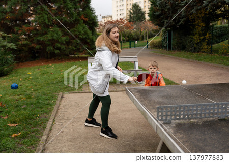 Mother playing outdoor table tennis pingpong with son in autumn park Mother playing outdoor table tennis pingpong with son in autumn park 137977883