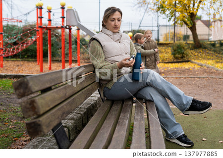 Mother relaxing on bench by playground while children play nearby at autumn 137977885