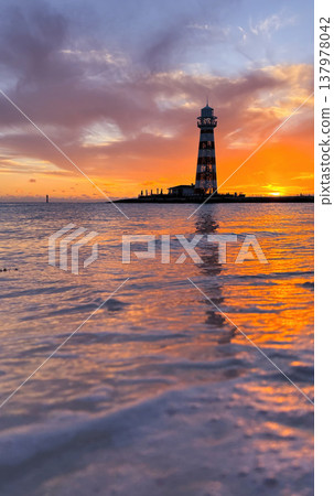 Vertical shot of a summer sunset. Sea foam in the foreground and the sun falling on the horizon with a lighthouse in the background. Vertical shot of a summer sunset. Sea foam in the foreground and the sun falling on the horizon with a lighthouse in the background. 137978042