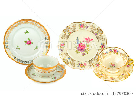 Elegant collection of vintage porcelain dinnerware featuring ornate gold rims and delicate floral patterns including tea cups and saucers isolated on a white background in horizontal view. Elegant collection of vintage porcelain dinnerware featuring ornate gold rims and delicate floral patterns including tea cups and saucers isolated on a white background in horizontal view. 137978309
