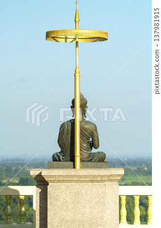 Rear view of a seated Buddha statue under a golden tiered umbrella on a high platform overlooking a lush landscape. Religious sculpture symbol of peace, meditation, and Buddhist spirituality. Rear view of a seated Buddha statue under a golden tiered umbrella on a high platform overlooking a lush landscape. Religious sculpture symbol of peace, meditation, and Buddhist spirituality. 137981915