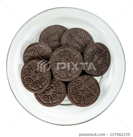 Top down view of a pile of dark chocolate sandwich cookies with floral patterns on a clear glass plate, isolated. Perfect for snack, dessert, or food industry marketing themes. 137982239