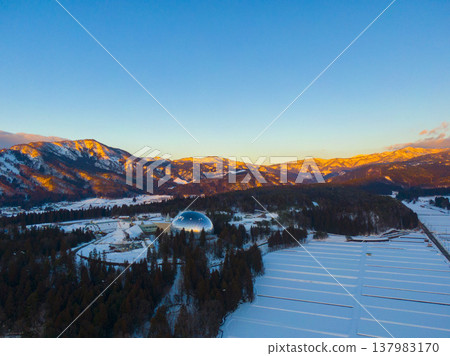 Snow-covered mountains bathed in the setting sun and the Fukui Prefectural Dinosaur Museum: Winter aerial photography by drone Snow-covered mountains bathed in the setting sun and the Fukui Prefectural Dinosaur Museum: Winter aerial photography by drone 137983170