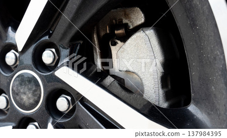 Close up shot focusing on a metallic silver brake caliper house behind a modern black and silver alloy wheel rim. Highlights the heavy duty texture of the caliper and the disc brake. Close up shot focusing on a metallic silver brake caliper house behind a modern black and silver alloy wheel rim. Highlights the heavy duty texture of the caliper and the disc brake. 137984395