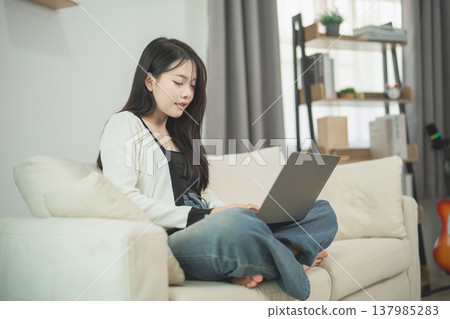 Young woman sitting on a cozy couch, using a laptop, focused on work or study, with a modern living room setting and soft natural light 137985283