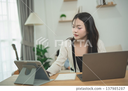 Young woman working from home with tablet and laptop in cozy modern workspace, focused and productive in bright interior during daytime Young woman working from home with tablet and laptop in cozy modern workspace, focused and productive in bright interior during daytime 137985287