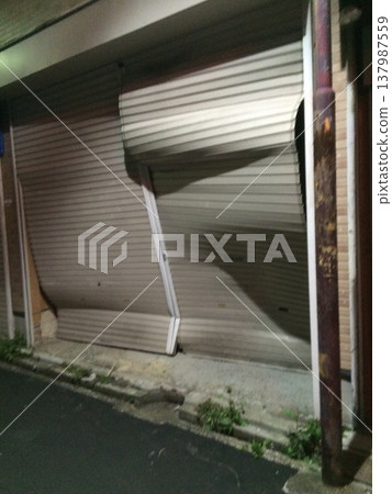 A scene of a heavily warped and broken metal shutter on a street corner at night. A scene of a heavily warped and broken metal shutter on a street corner at night. 137987559