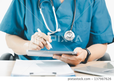 Medical professional in blue scrubs using tablet and stylus with stethoscope, documenting patient records at clinic desk, concept of digital health, telemedicine and modern healthcare workflow. Medical professional in blue scrubs using tablet and stylus with stethoscope, documenting patient records at clinic desk, concept of digital health, telemedicine and modern healthcare workflow. 137987626