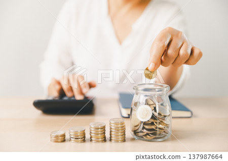 Hand dropping coin into glass savings jar on desk with scattered coins, notebook and phone. Budget planning, personal finance, frugal living, investment goals, future security. investment goals Hand dropping coin into glass savings jar on desk with scattered coins, notebook and phone. Budget planning, personal finance, frugal living, investment goals, future security. investment goals 137987664