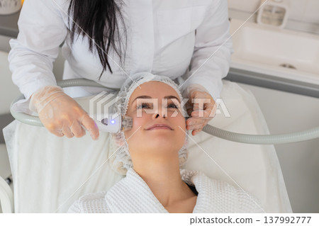 Woman receives RF lifting facial massage therapy by cosmetologist using professional equipment Woman receives RF lifting facial massage therapy by cosmetologist using professional equipment 137992777