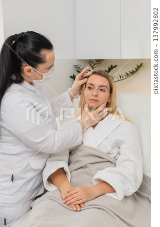 Cosmetologist in mask checking facial skin of young woman client before cosmetic treatment in clinic 137992802
