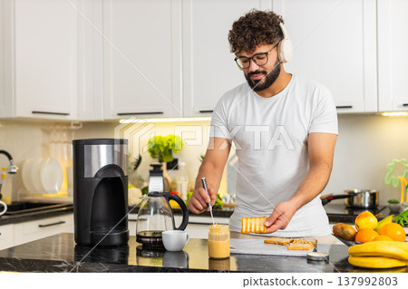 Arabian young adult man dancing with headphones spreading peanut butter toast breakfast in kitchen Arabian young adult man dancing with headphones spreading peanut butter toast breakfast in kitchen 137992803