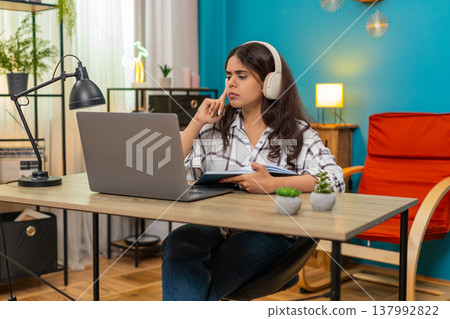 Indian woman wearing headphones nods during online lecture on laptop writing notes in notebook study Indian woman wearing headphones nods during online lecture on laptop writing notes in notebook study 137992822