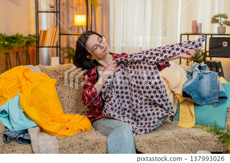 Young woman sorting clothes on home sofa, thoughtfully deciding what to keep and what to give away Young woman sorting clothes on home sofa, thoughtfully deciding what to keep and what to give away 137993026