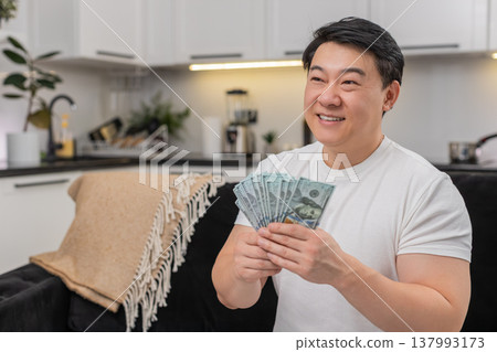 Asian man counts cash dollars smiles makes money sign and plans invest purchase bonus salary savings Asian man counts cash dollars smiles makes money sign and plans invest purchase bonus salary savings 137993173