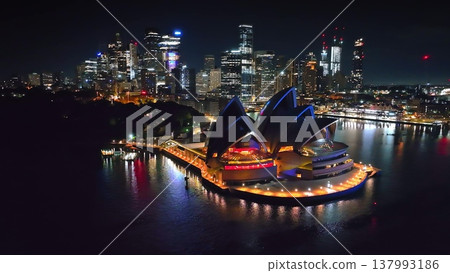 Sydney Opera House aerial panorama at night. Modern building stands prominently with its iconic sails lit against dark harbor water, overlooked by vibrant illuminated city lights skyline in Australia Sydney Opera House aerial panorama at night. Modern building stands prominently with its iconic sails lit against dark harbor water, overlooked by vibrant illuminated city lights skyline in Australia 137993186