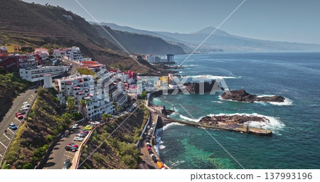 Tenerife: Aerial drone flight over colorful hotel resort complex buildings of coastal town cityscape on hill, pier, deep blue Atlantic Ocean, with the distant Pico del Teide volcano in background Tenerife: Aerial drone flight over colorful hotel resort complex buildings of coastal town cityscape on hill, pier, deep blue Atlantic Ocean, with the distant Pico del Teide volcano in background 137993196