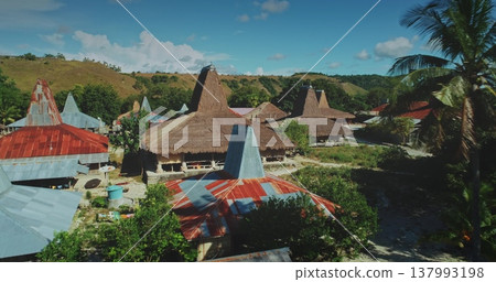 Aerial view of a village of traditional houses with conical roofs made of straw and metal sheets. Surrounded by green vegetation and hills in Sumba Island. Indonesia. On a sunny day with some clouds 137993198