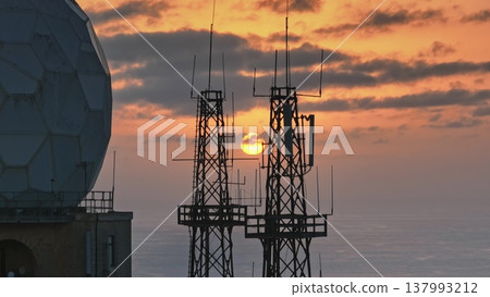 Telecommunication towers and radar dome silhouette against a vibrant orange and yellow sunset over a vast ocean, creating a striking contrast between technology and nature Telecommunication towers and radar dome silhouette against a vibrant orange and yellow sunset over a vast ocean, creating a striking contrast between technology and nature 137993212