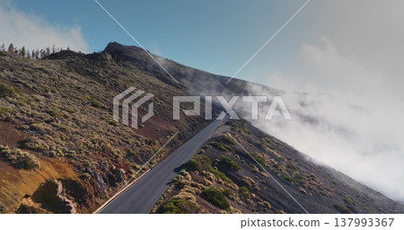 Tenerife North: mountain road cutting across the rugged volcanic hill terrain, with low lying clouds drifting along the slopes under a clear blue sky. Aerial view drone flight 137993367