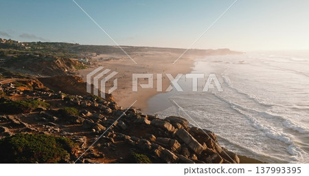 Portugal, Lisbon: Aerial view Praia do Guincho beach coastline with ocean waves rolling yellow sand, people relax and enjoy warm golden hour light sunset. Drone flight over rocky cliffs, zoom out 137993395