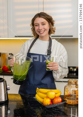 Young woman making green smoothie in home kitchen smiling during healthy diet breakfast ritual plan 137994146